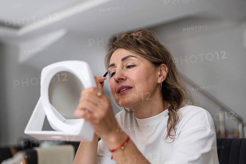 Portrait of mature woman holding mirror and tweezers. woman is focused on applying makeup in her home. She holds a small tool with one hand while looking into a mirror