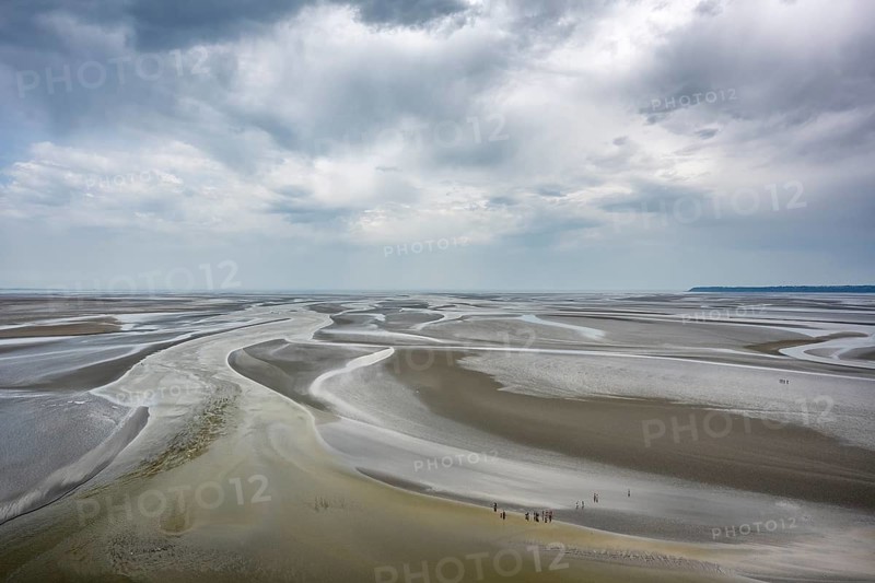 View from the rocky island, monastery island of Le Mont Saint Michel onto a wide mudflat landscape with creeks and dramatic clouds at low tide, Mont-Saint-Michel Bay, Pontorson, Manche department, Normandy, France