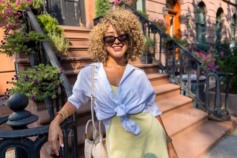 Happy young woman with curly hair wearing sunglasses, light shirt, and yellow dress, smiling while leaning on a railing in front of new york city brownstone steps on a sunny day