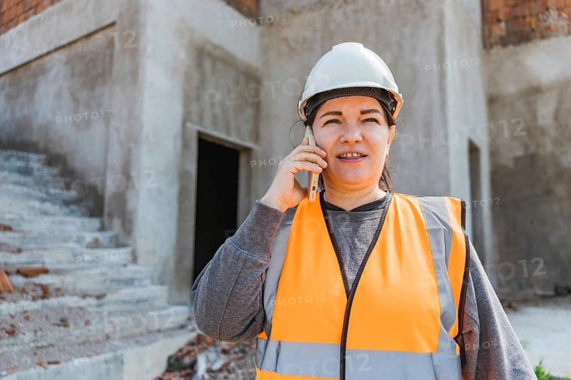 Woman in a construction site wearing a hard hat and orange safety vest. She is talking on a mobile phone with a serious expression