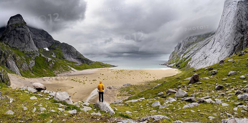 Young man in front of sandy beaches between rocky mountain peaks and dramatic clouds, Lofoten, Norway