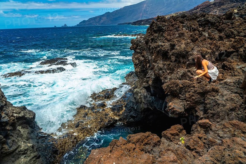 Woman crouching on dark volcanic rocks, observing the vibrant turquoise ocean water crashing against the rugged coastline of el hierro in the canary islands