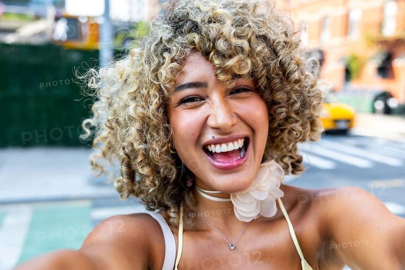 Young woman with curly blonde hair smiling broadly and taking a selfie, enjoying an urban lifestyle experience in a new york city street with a yellow cab in the background