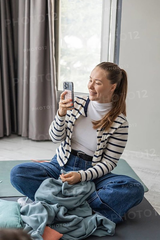 A young Caucasian woman with long brown hair sits cross-legged on a mat, smiling while holding a smartphone. She wears a striped cardigan and jeans, enjoying a live music concert at home