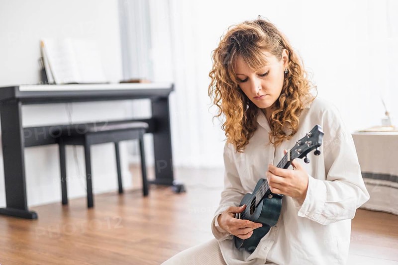 Person sits with a ukulele near a piano in a room with wooden floors