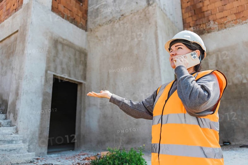 Woman in a hard hat and safety vest talks on her phone at a construction site. The background features a partially built structure and greenery