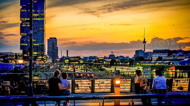 Romantic scene, people sitting on bridge looking at sunset, EDGE East Side high-rise building, above the skyline with television tower, view from Modersohn Bridge, Friedrichshain, Berlin