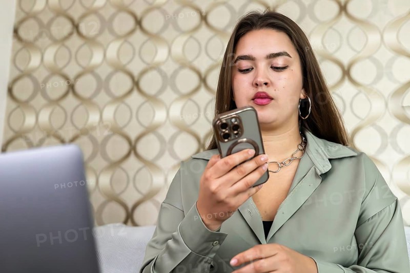 A young woman sits on a couch and looks at her smartphone. She appears focused while a laptop is placed on a table nearby. The setting has a light background design