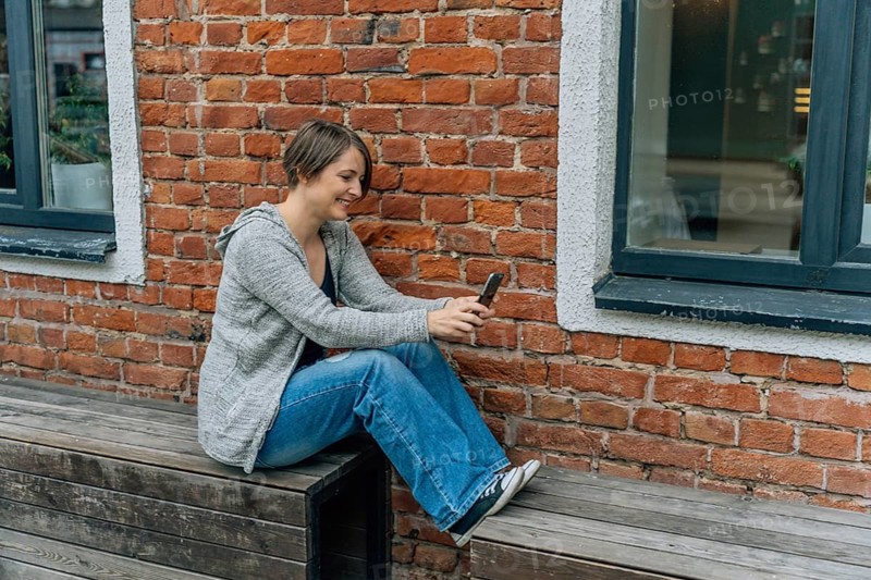 Smiling Woman Using Smartphone on a Wooden Bench, A woman casually sits on a bench using her smartphone against a brick wall