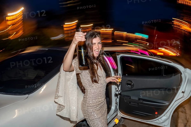 Young woman in a glamorous dress joyfully exiting a white limousine, raising a champagne bottle and glass, celebrating a special occasion with motion blur indicating a lively nightlife experience