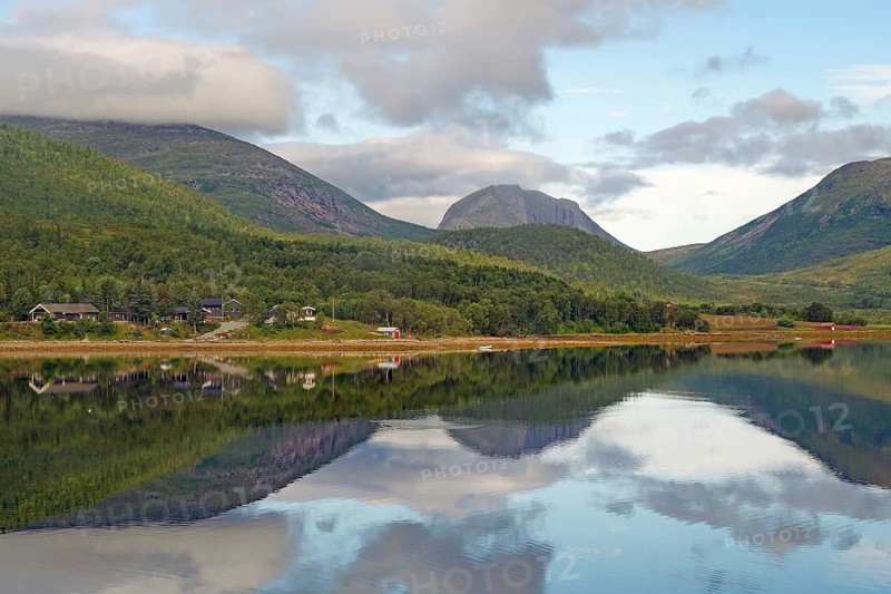 Landscape with mountains and a lake with calm reflection, surrounded by forest and small huts under a cloudy sky, Fjordbotn Camping, Botnhamm, Troms, Senja, Norway