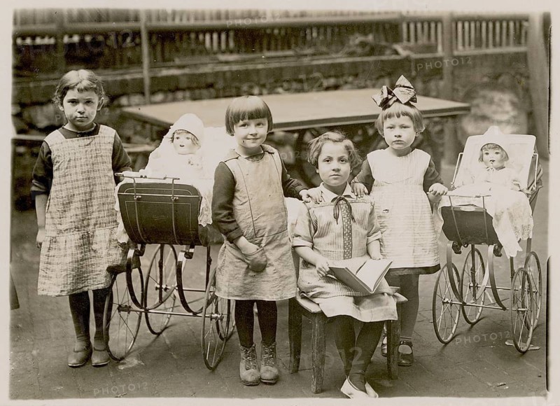Five girls posing with dolls and doll prams in a backyard, schoolbook, children, around 1920
