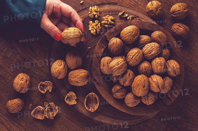 A woman's hand taking a walnut from a wooden bowl filled with walnuts. There are shells and walnuts scattered around