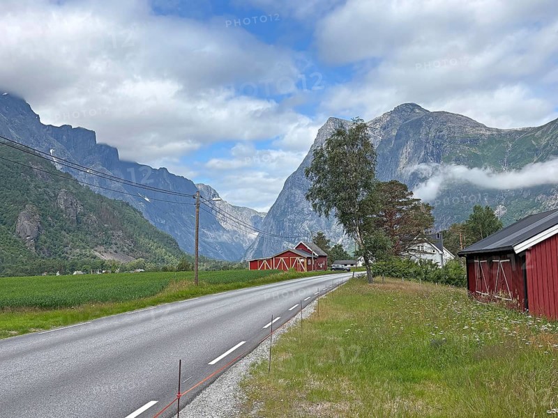 A road leads through a green valley flanked by mountains and red houses under a partly cloudy sky, Romsdalshornet, Raumadalen, Trollvegen, Andalsnes, Norway