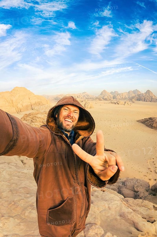 Smiling male tourist traveler wearing hooded traditional brown farwa cloak taking selfie making victory finger gesture vast sand desert valley mountain landscape backdrop Wadi Rum Jordan