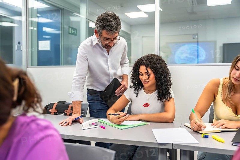 University professor mentoring a diverse female student over a document in a modern classroom while peers observe and engage in focused academic discussion and collaboration