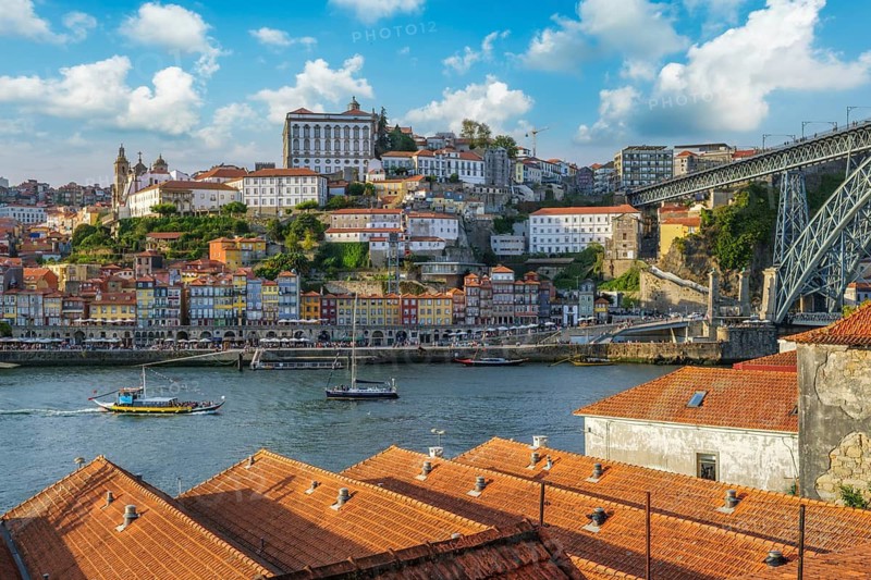 View of Porto Ribeira, Portugal, across Douro River over house rooftops, with Dom Luis I Bridge, river boats, and orange rooftops in the foreground