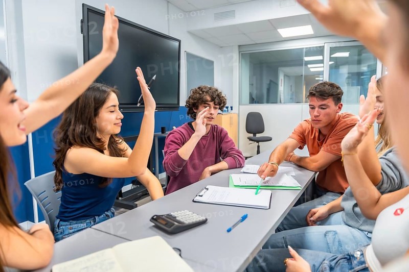 Diverse students actively participating in a classroom discussion, raising hands, and sharing ideas while working together at a table, highlighting concepts of learning, teamwork, and engagement