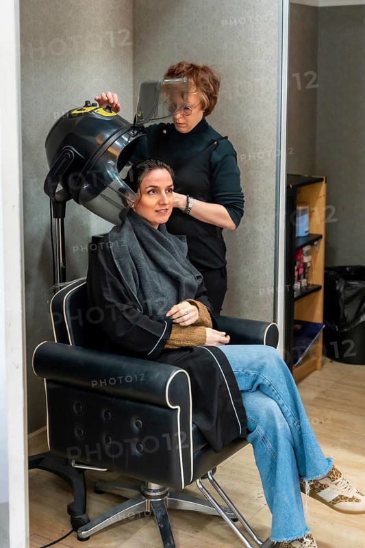 Professional hairdresser providing a hair treatment service to a woman sitting comfortably in a salon chair, using a hooded dryer for her hair, showcasing beauty and personal care concepts