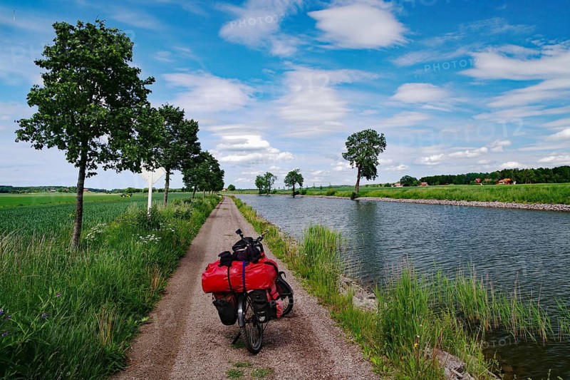 A loaded bicycle stands on a narrow path along a river under slightly cloudy sky, bicycle tours, cycle tourism, Göta Canal, Töreboda, Västra Götalands län, Sweden