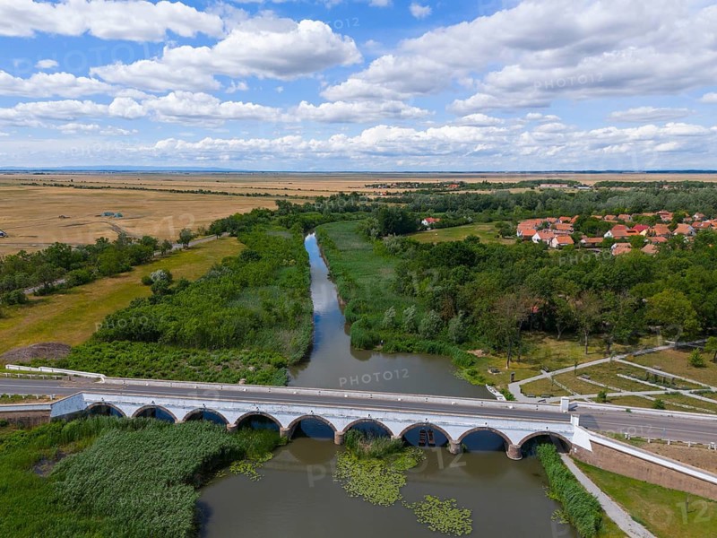 Wide landscape with a river and a bridge surrounded by fields and a village under a cloudy sky, aerial view, nine-arched stone bridge, Kilenclyukú híd, Hortobagy, Hortobagy National Park, Hortobágyi Nemzeti Park, Hortobágyi Csillagoségbolt Park, Puszta, steppe area, UNESCO biosphere reserve, Hungary