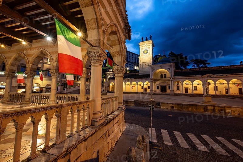 Arcades of the Loggia del Lionello with Italian flags, illuminated main square Piazza della Libertà with Loggia e Tempietto di San Giovanni and clock tower, Castello di Udine behind, blue hour, dramatic sky, old town, Udine, Friuli-Venezia Giulia, Italy