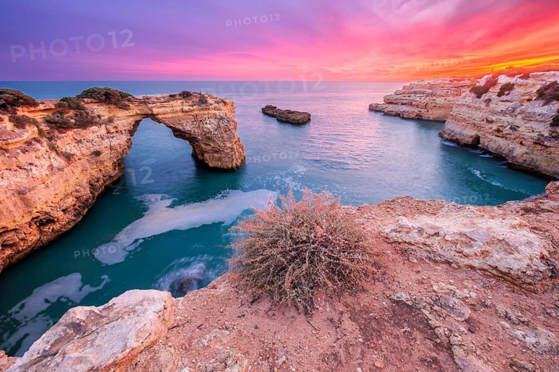 Dramatic sunset view of Arco de Albandeira at Praia de Albandeira in Algarve, Portugal, with pink sky, limestone sea arch, Atlantic water, and a high Atlantic view over the cliff edge