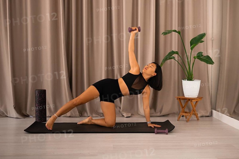 Woman practicing a yoga pose on a workout mat with a plant and dumbbells nearby