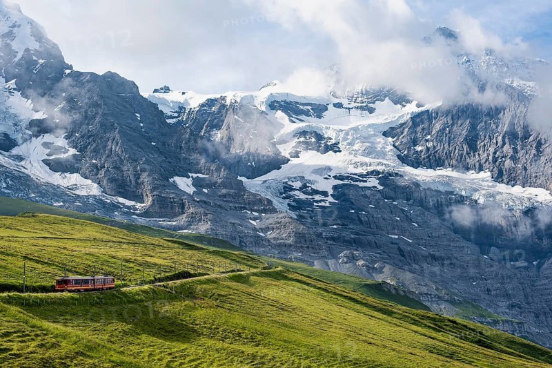 Jungfrau Railway to Jungfraujoch runs through green meadows, above the Kleine Scheidegg stop, rack railway, in the back mountains with peaks Eiger and Mönch, Jungfrau region, Bernese Oberland, Switzerland