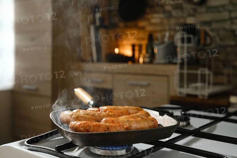 Banger sausages frying in cast-iron griddle pan on stovetop in kitchen. Smoke from melted fat rises above skillet