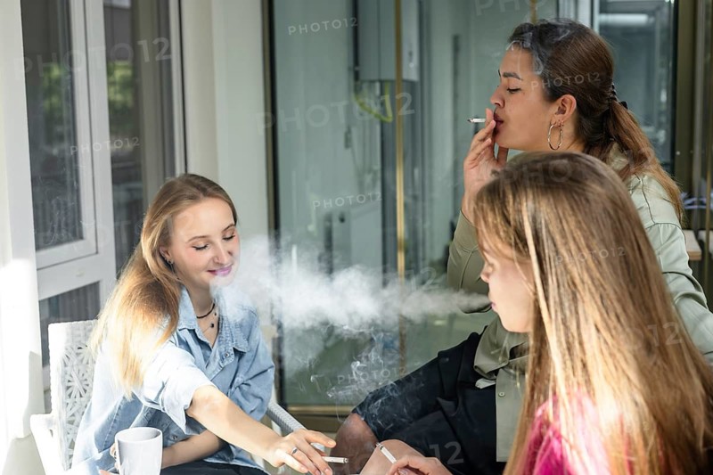 Three friends are sitting at a table in a cafe during the afternoon. They are smoking cigarettes and chatting while enjoying their time. One friend is reaching for a cigarette