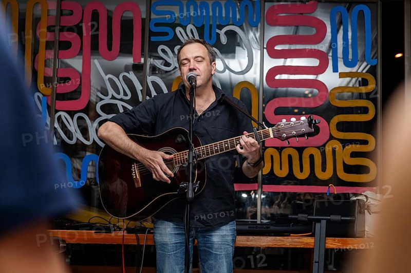 A middle-aged Caucasian man with short hair sings into a microphone while playing an acoustic guitar. Colorful background with abstract designs