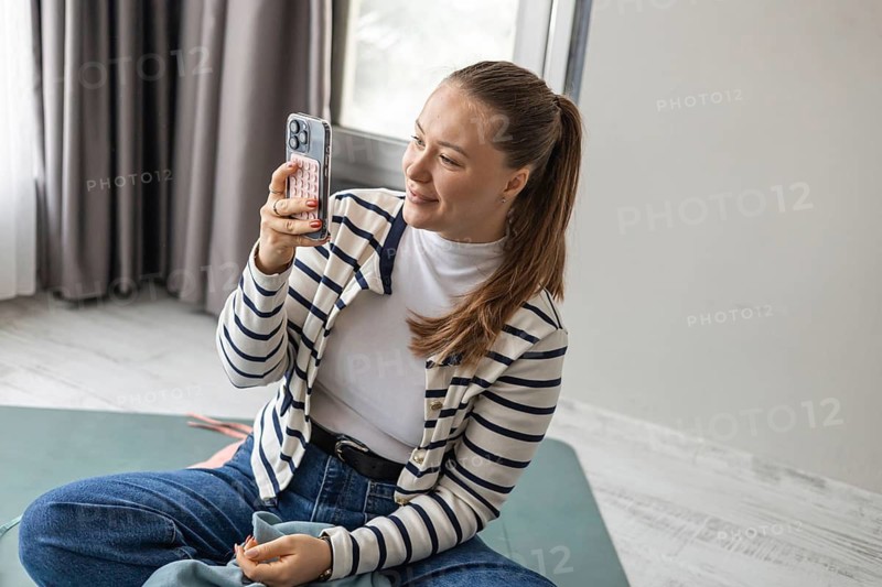 A young Caucasian woman with long brown hair sits cross-legged on a mat, smiling while holding a smartphone. She wears a striped cardigan and jeans, enjoying a live music concert at home