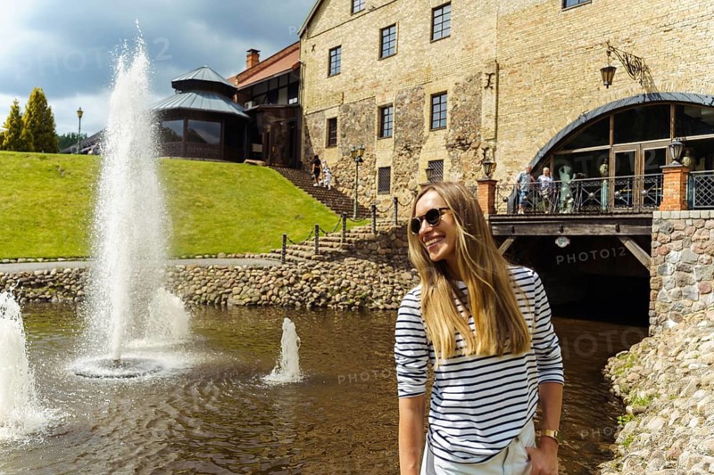 A long-haired woman in a striped jumper and sunglasses laughs next to a fountain in the summer, A woman smiling joyfully near a fountain with a historic building in the background on a sunny day, Belmontas Park, Vilnius, Lithuania