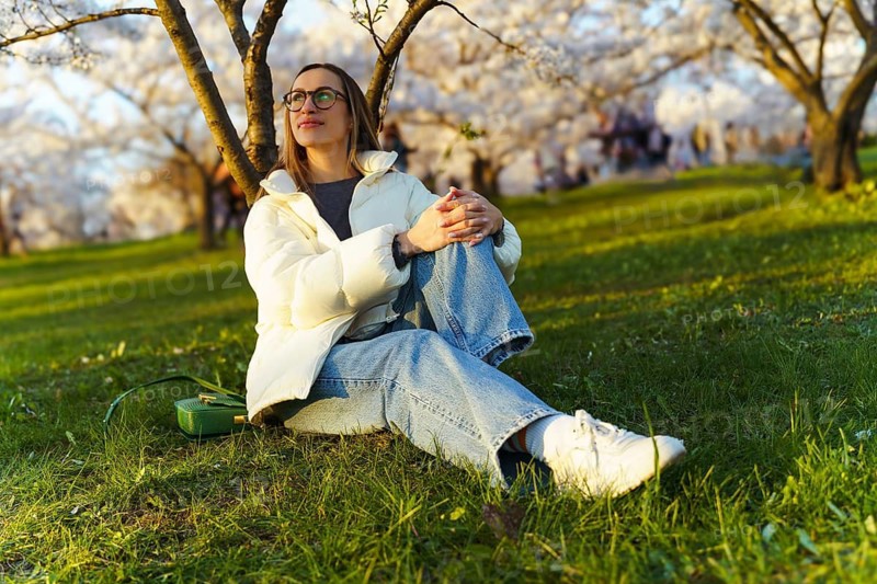 Young woman sitting on grass in blooming cherry garden and looking at sakura trees, Woman sitting on grass in a park surrounded by blossoming trees, looking relaxed and content
