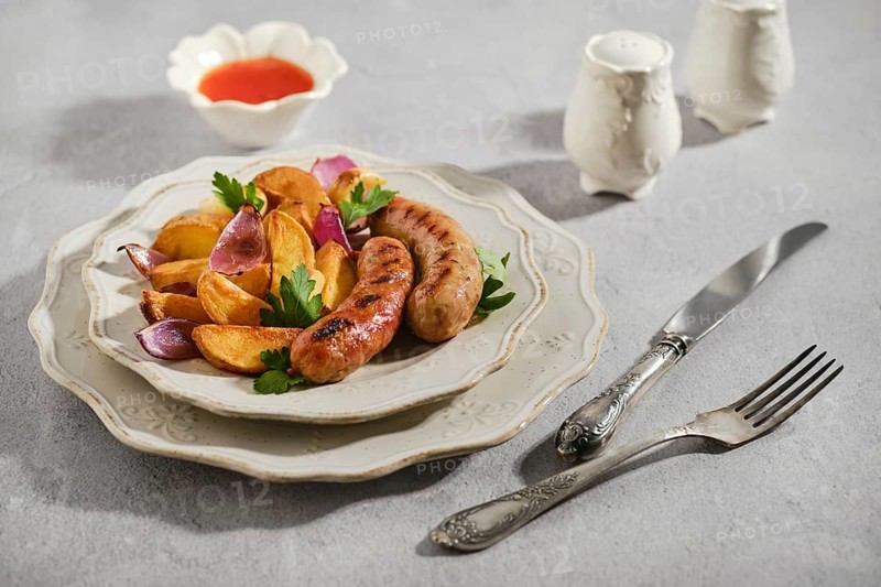 Grilled banger sausages with roasted potatoes, red onion and herbs. Small bowl with sauce sits nearby. Setting shows grey tabletop with cutlery arranged beside plate