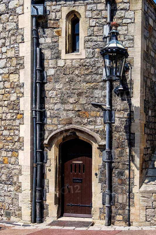 A small arched wooden doorway sits beside a mullioned window on a rubble stone wall at Windsor Castle, flanked by weathered cast iron drainpipes and an overhanging lantern bracket that speak to the castle's practical working fabric, Windsor, England, United Kingdom