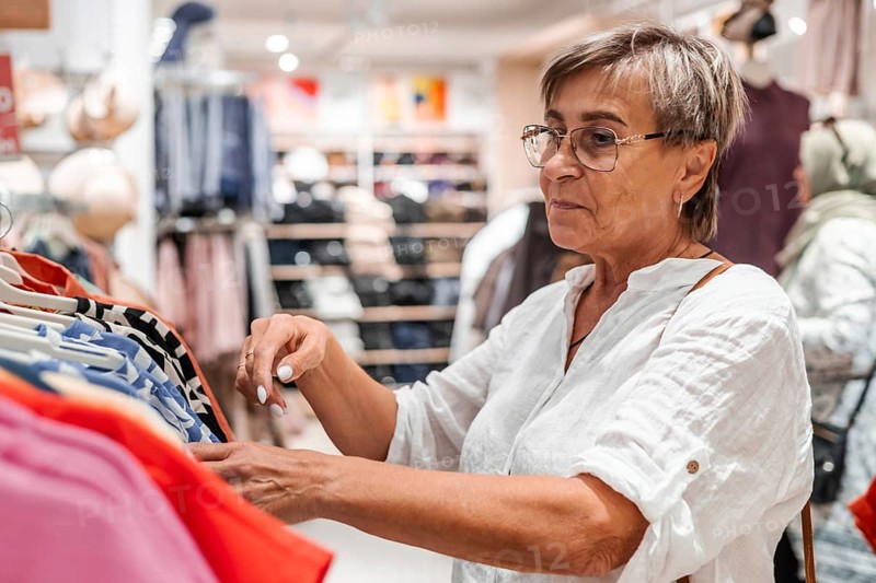 Senior Caucasian woman with short gray hair shopping for clothes in a retail store. She is examining a colorful shirt while browsing through a rack of clothing
