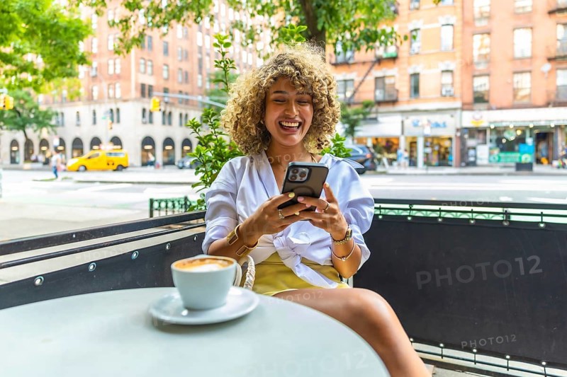 Young woman with curly hair laughing and using smartphone while sitting at an outdoor cafe table with a coffee, enjoying urban life on a manhattan street