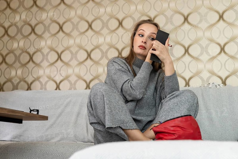 A young Caucasian woman with long brown hair applies makeup while sitting on a couch at home. She wears a gray outfit and looks focused in a handheld mirror. Home daily beauty routine
