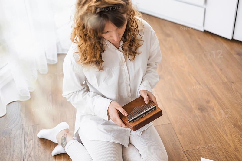 A woman in white seated on the wooden floor, playing a kalimba