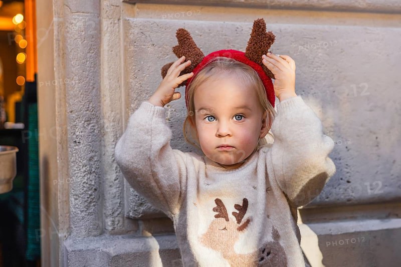 A young Caucasian girl with light brown hair wears a reindeer headband and a cozy sweater. She stands against a textured wall, looking serious and festive