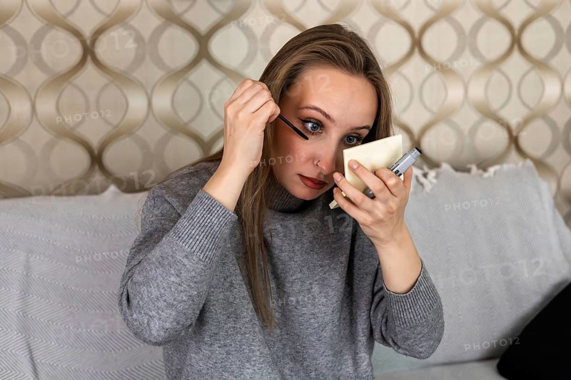 Young Caucasian woman with long brown hair applies makeup in a cozy home setting. She sits on a couch, holding a mirror and a makeup brush