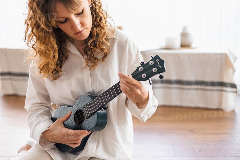 Focused individual in white shirt playing ukulele on a wooden floor