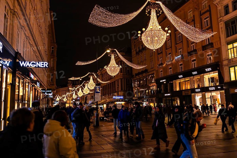Vienna, Austria - 15 December, 2025: Busy street in Vienna, Austria, illuminated by festive lights. Shimmering chandeliers hang above shops. People of diverse backgrounds walk in winter attire