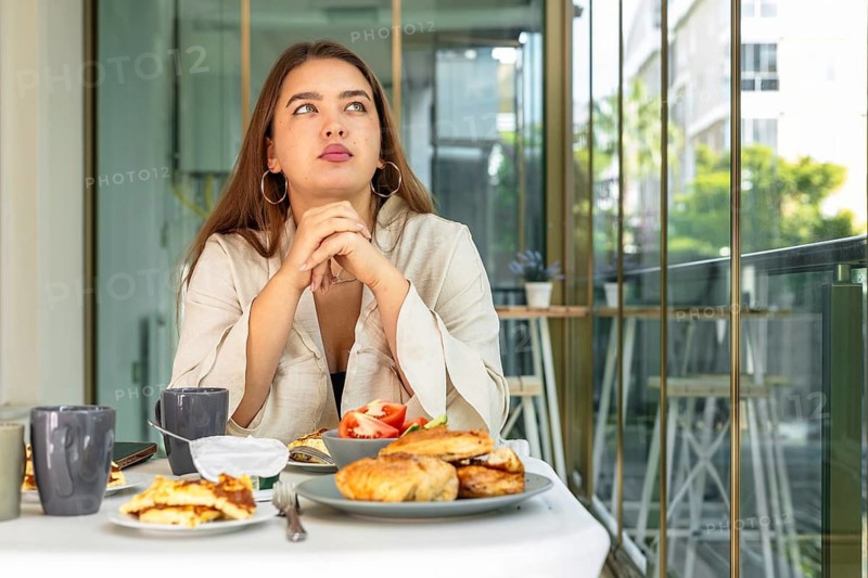 A young Hispanic woman with long brown hair sits at a table, looking thoughtfully at a spread of breakfast foods, including pastries and fruit, in a bright, modern setting