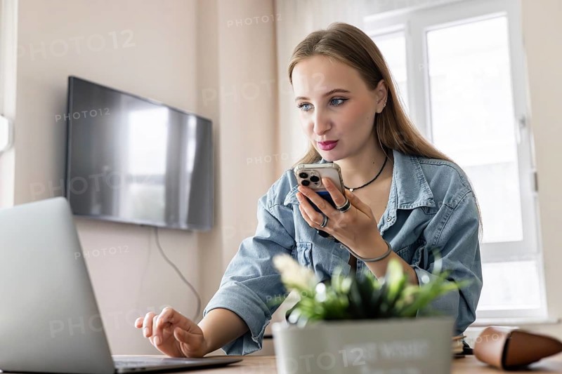 Young Caucasian woman with long blond hair using a smartphone while working on a laptop at a desk. Indoor setting with a plant in the foreground