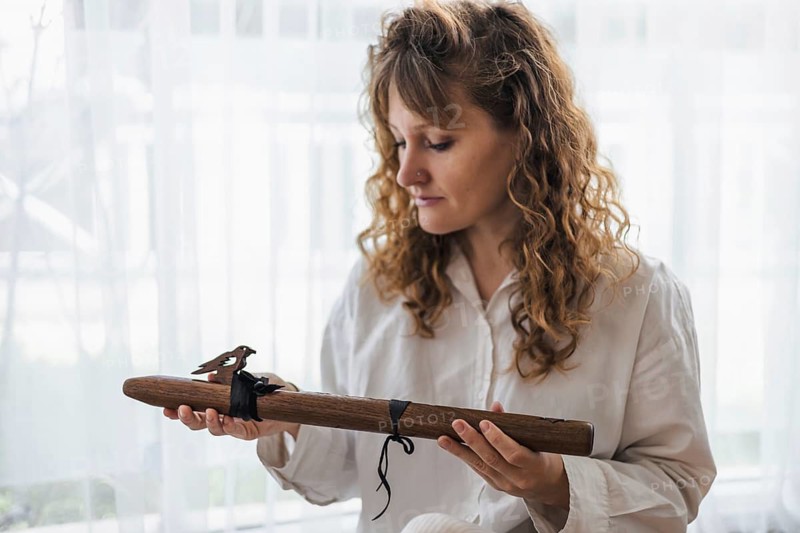 A woman contemplatively holds a wooden flute, seated by a softly lit window