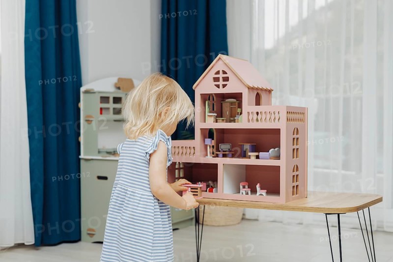 Child in a blue dress playing with a dollhouse set on a wooden table in a bright room