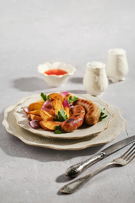 Grilled sausages rest on white plate beside colorful roasted vegetables. Small dish of sauce sits nearby. Fork and knife lie next to plate. Meal is set on light surface
